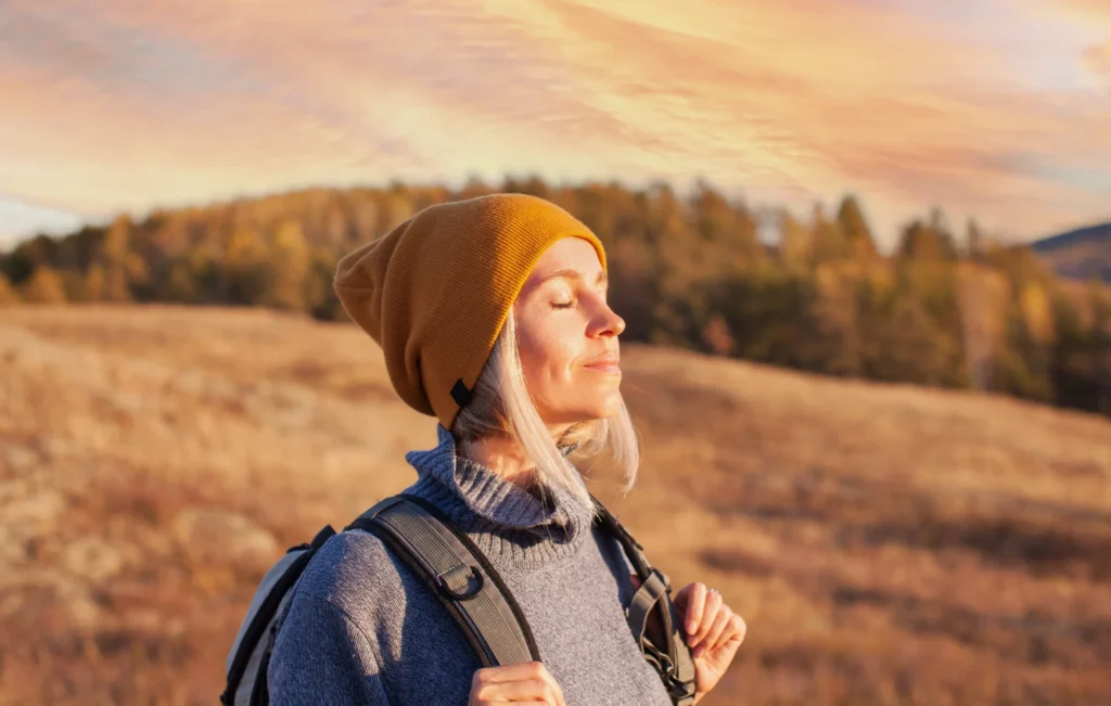 Woman enjoying mindful breathing outdoors at sunrise, wearing a warm hat and backpack in a peaceful autumn landscape. Symbolising relaxation, rejuvenation, and connection with nature — reflecting Re:nu’s holistic wellness and recovery philosophy.