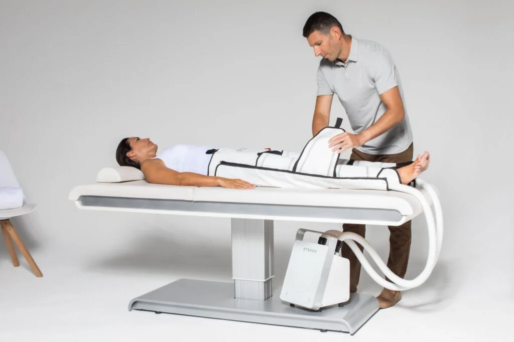 Male wellness specialist fitting the Stendo compression suit around a woman's legs as she lies on a treatment bed