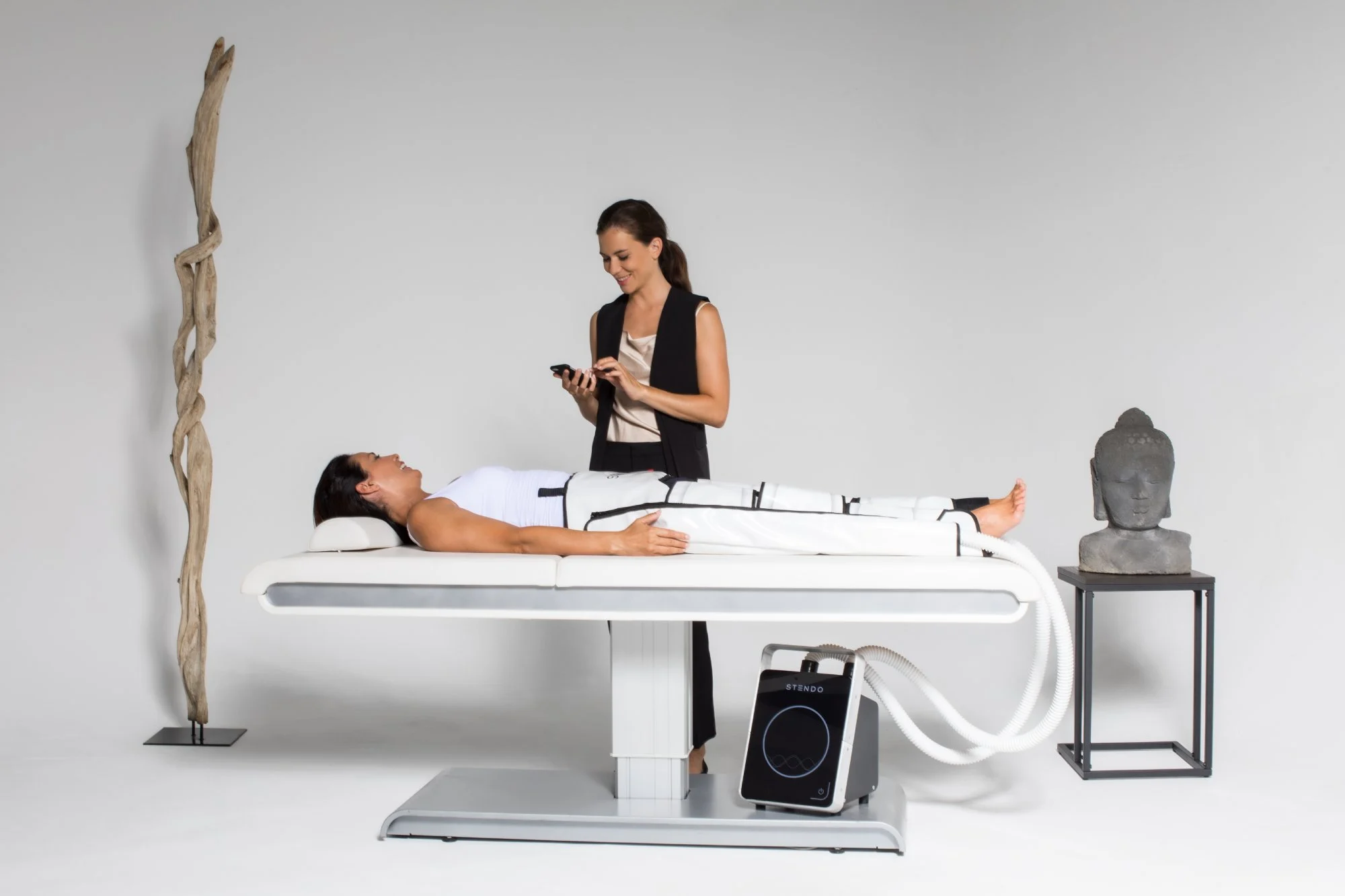 A woman lying down on a treatment bed wearing the Stendo compression suit as a wellness specialist monitors the treatment device